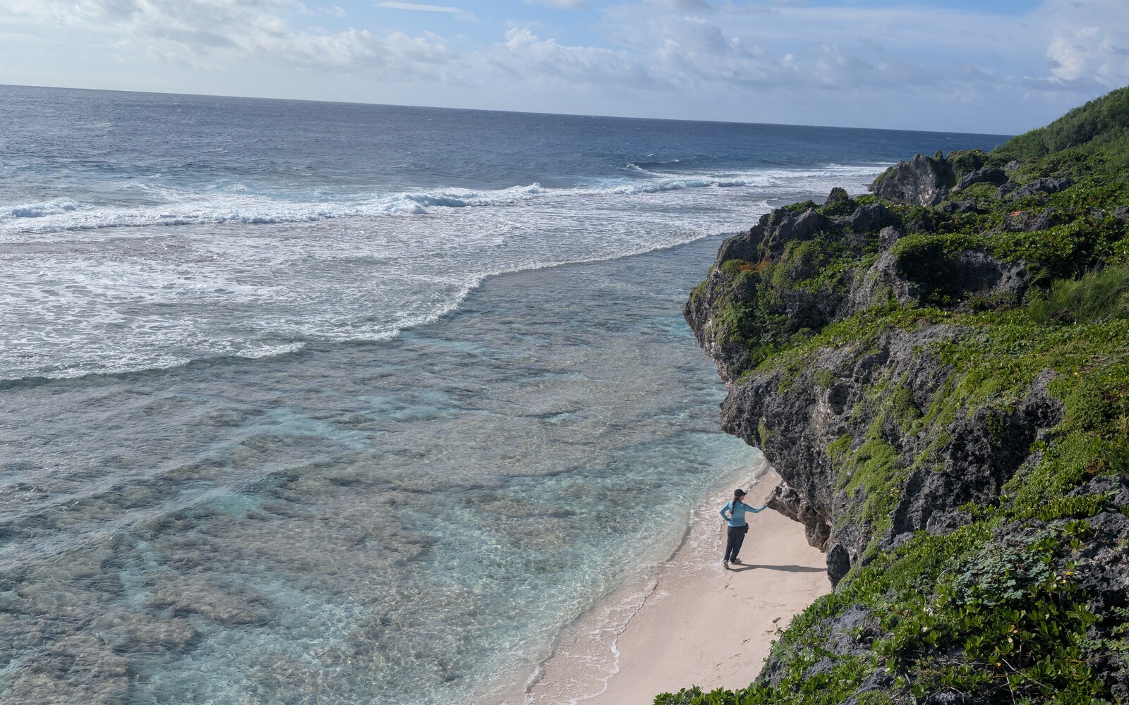 Oana standing on a narrow beach beneath a dark, vegetation-covered limestone overhang, beside shallow turquoise reef water and breaking waves at Tarapaku, Atiu.