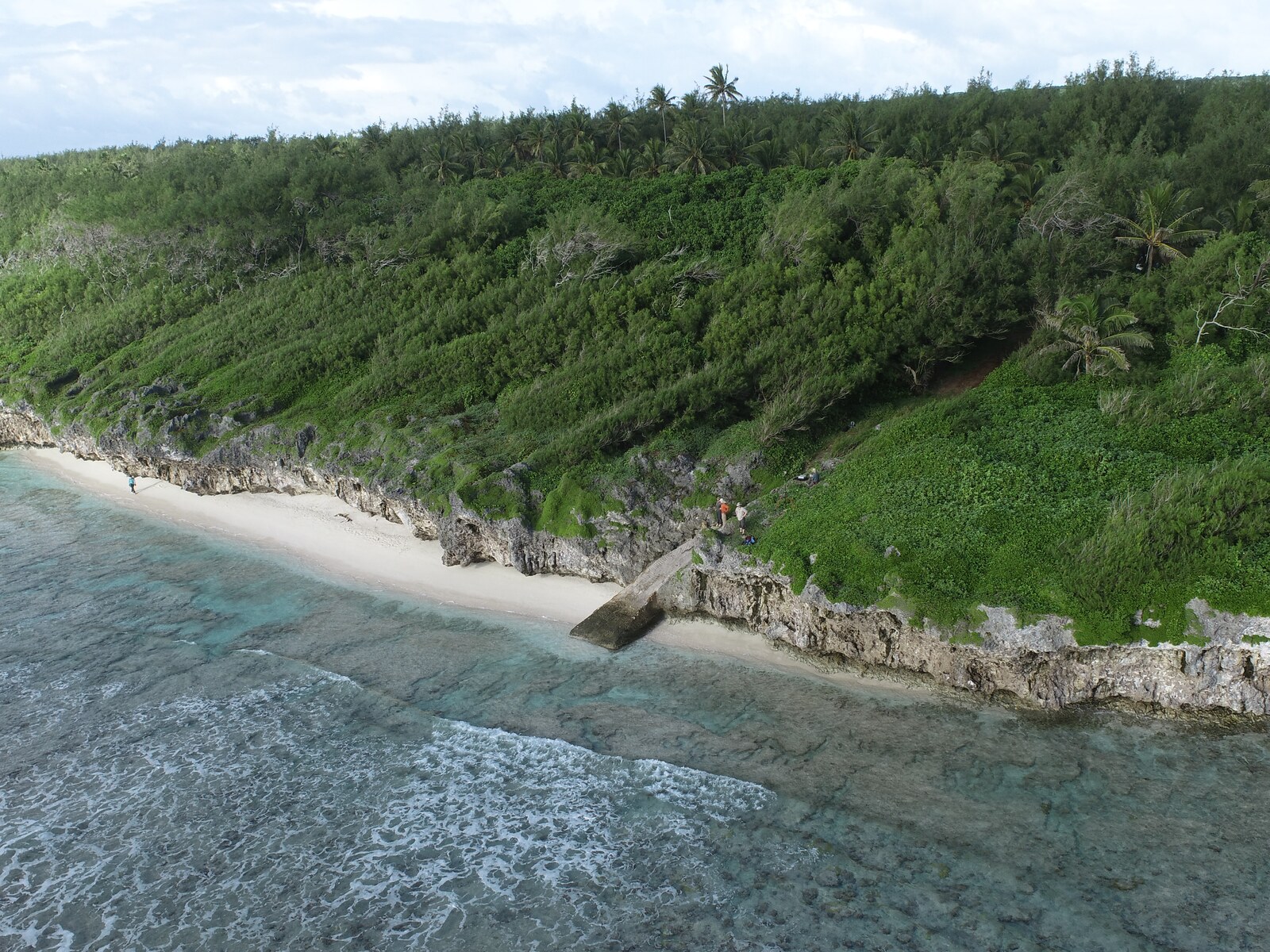Low-oblique drone view of HS106 near Tarapaku, with fieldworkers along a green limestone coast above a narrow pale beach, shallow turquoise reef flat, and breaking waves.