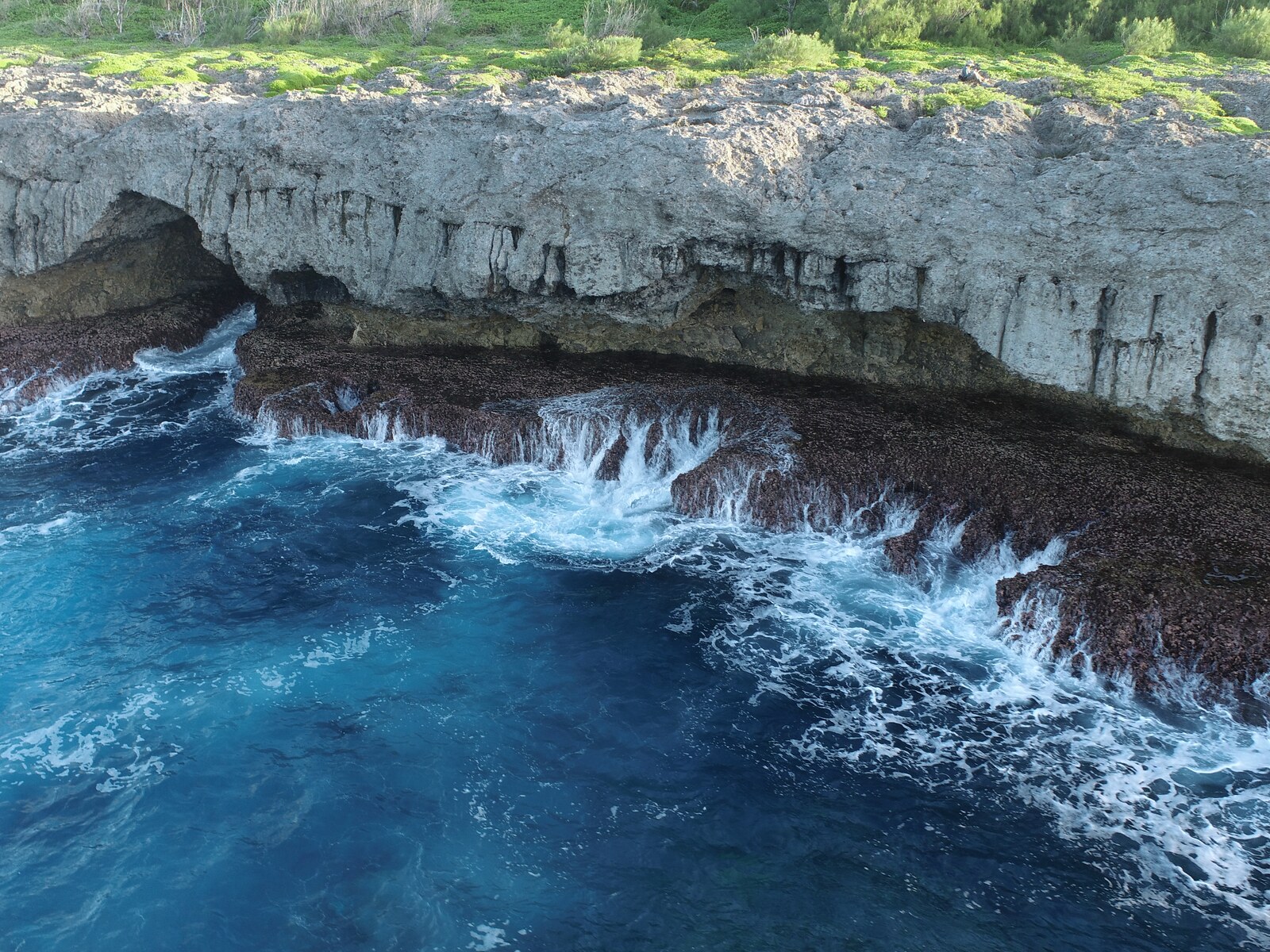 Low-oblique drone view at HS107 near Totiko, with turquoise waves spilling across a dark coastal bench below a pale undercut limestone cliff and rough ground with low vegetation.
