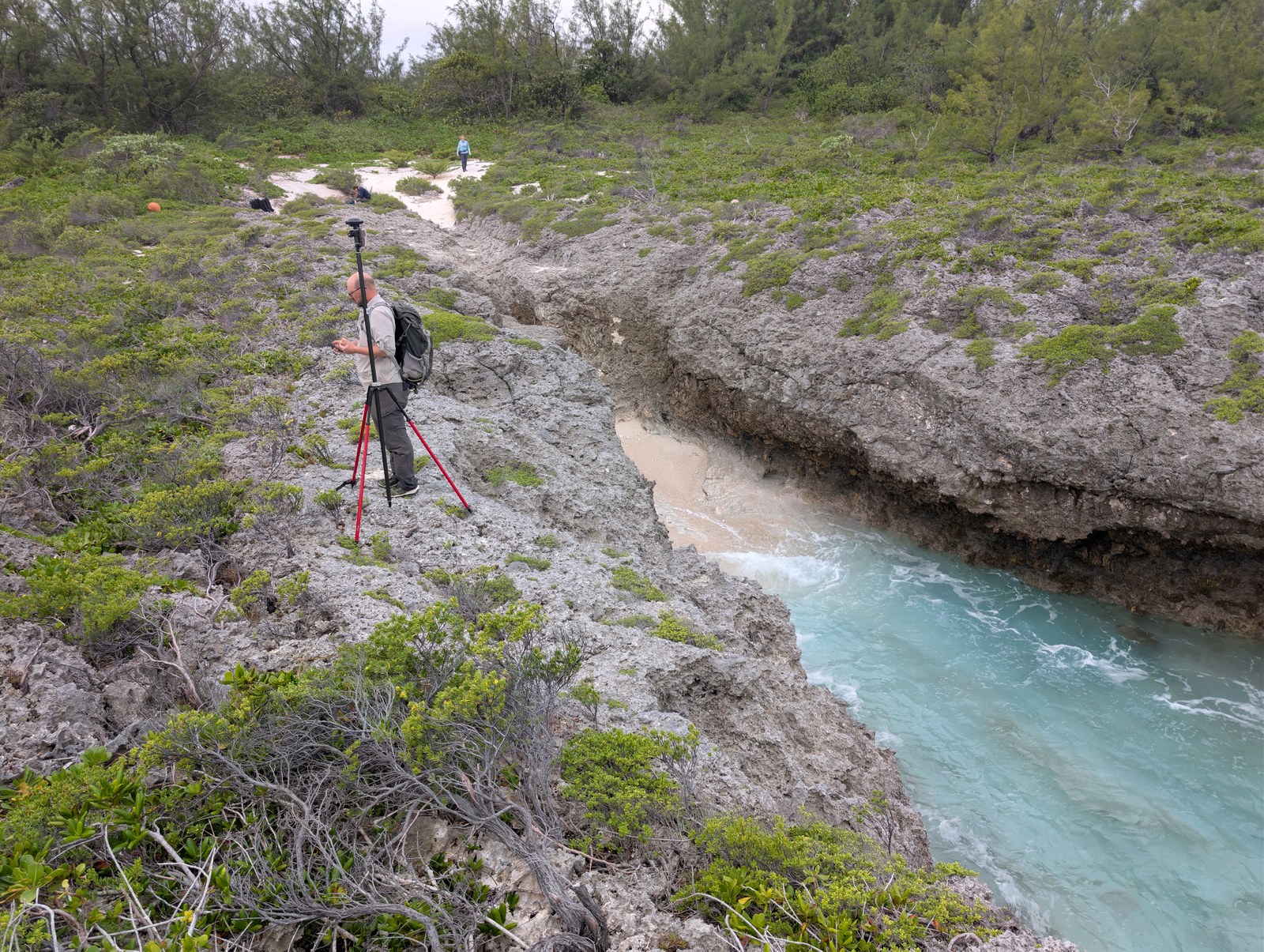 Two fieldworkers among jagged reef rock at HS124, with one beside a red survey tripod above a narrow turquoise channel and the other near a pale inland track.