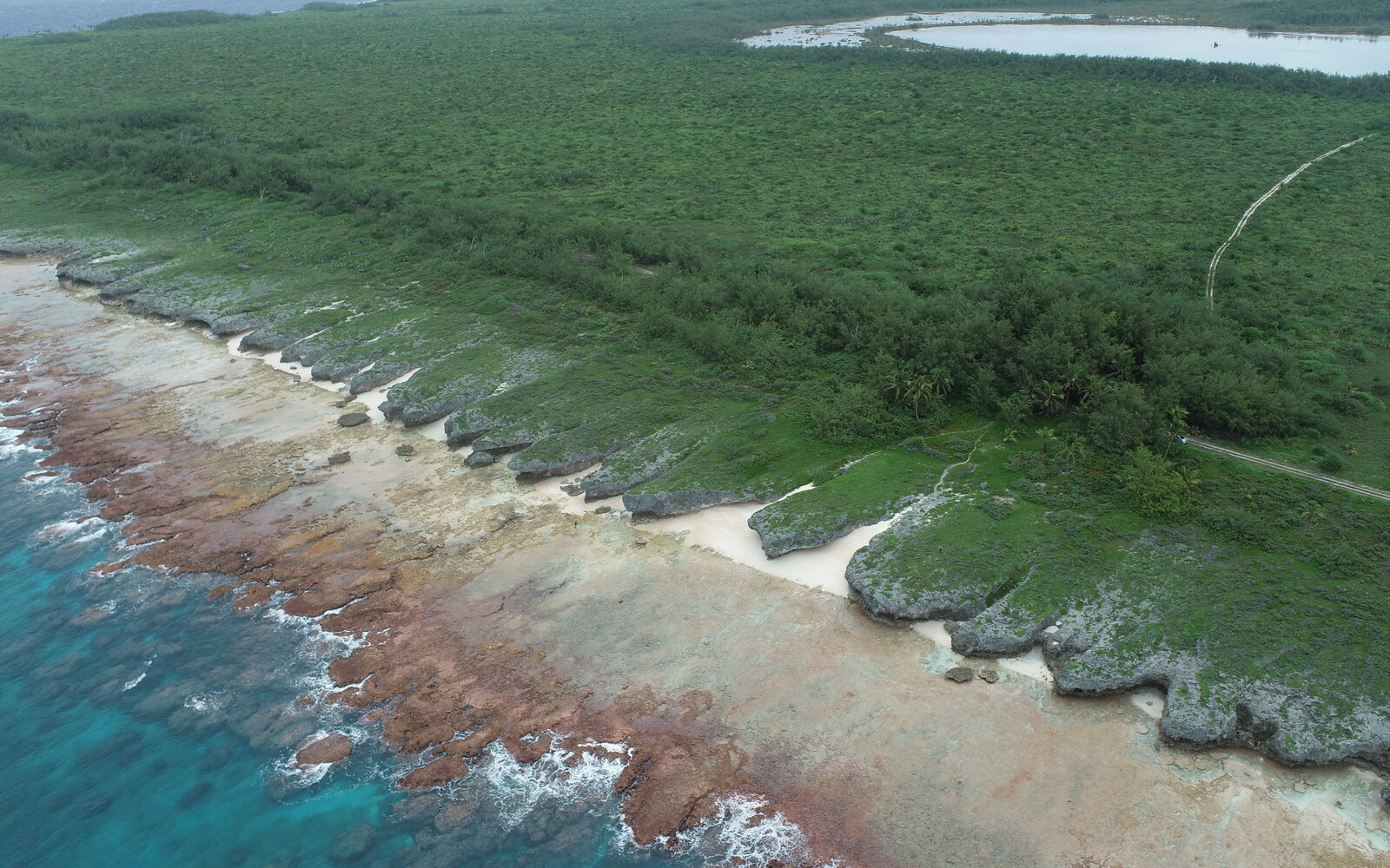 Low-oblique drone view across Te Rotonui’s wave-washed reef flat and repeated coastal limestone spurs toward dense vegetation, an access track, and an inland lake on Mitiaro.