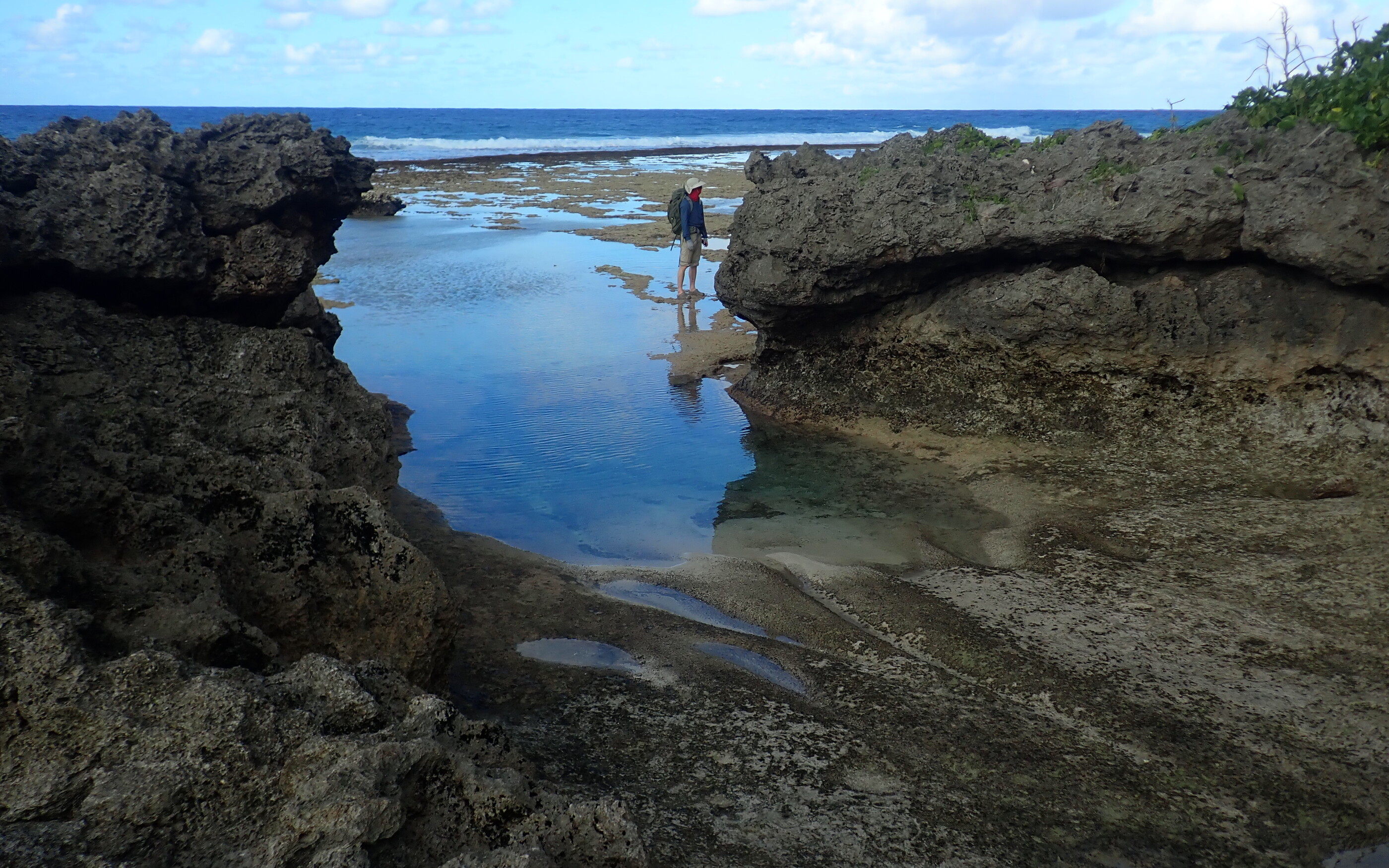 Roger standing on the exposed reef surface beside a blue pool between dark weathered coastal rocks, with the reef flat, breaking waves, and open ocean beyond at Ngatangiia, Rarotonga.