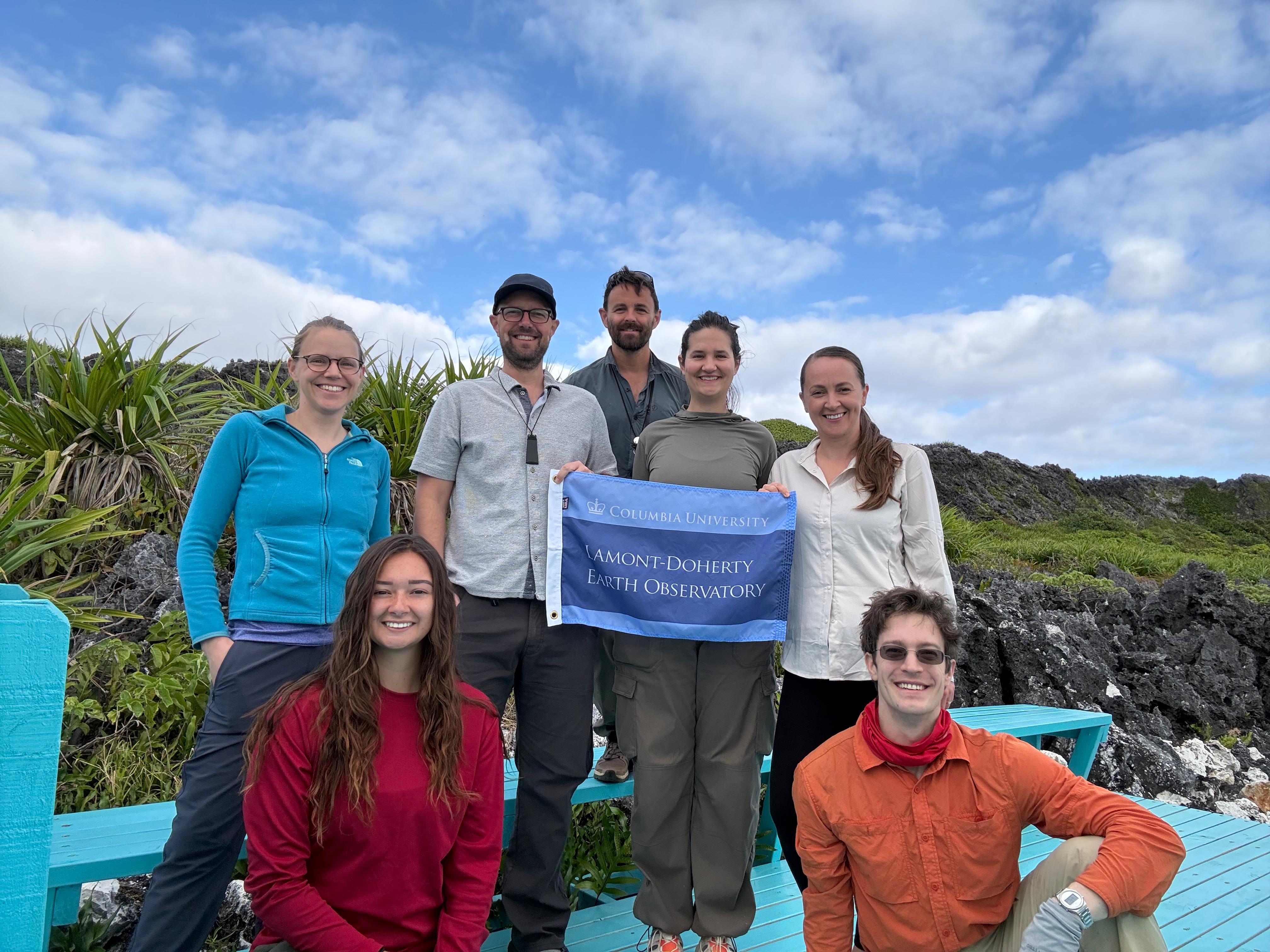 Seven members of the Cook Islands 2026 HISEAS field team stand together on a turquoise bench amid dark raised-reef limestone and coastal plants on Mangaia, holding a Lamont-Doherty Earth Observatory flag.